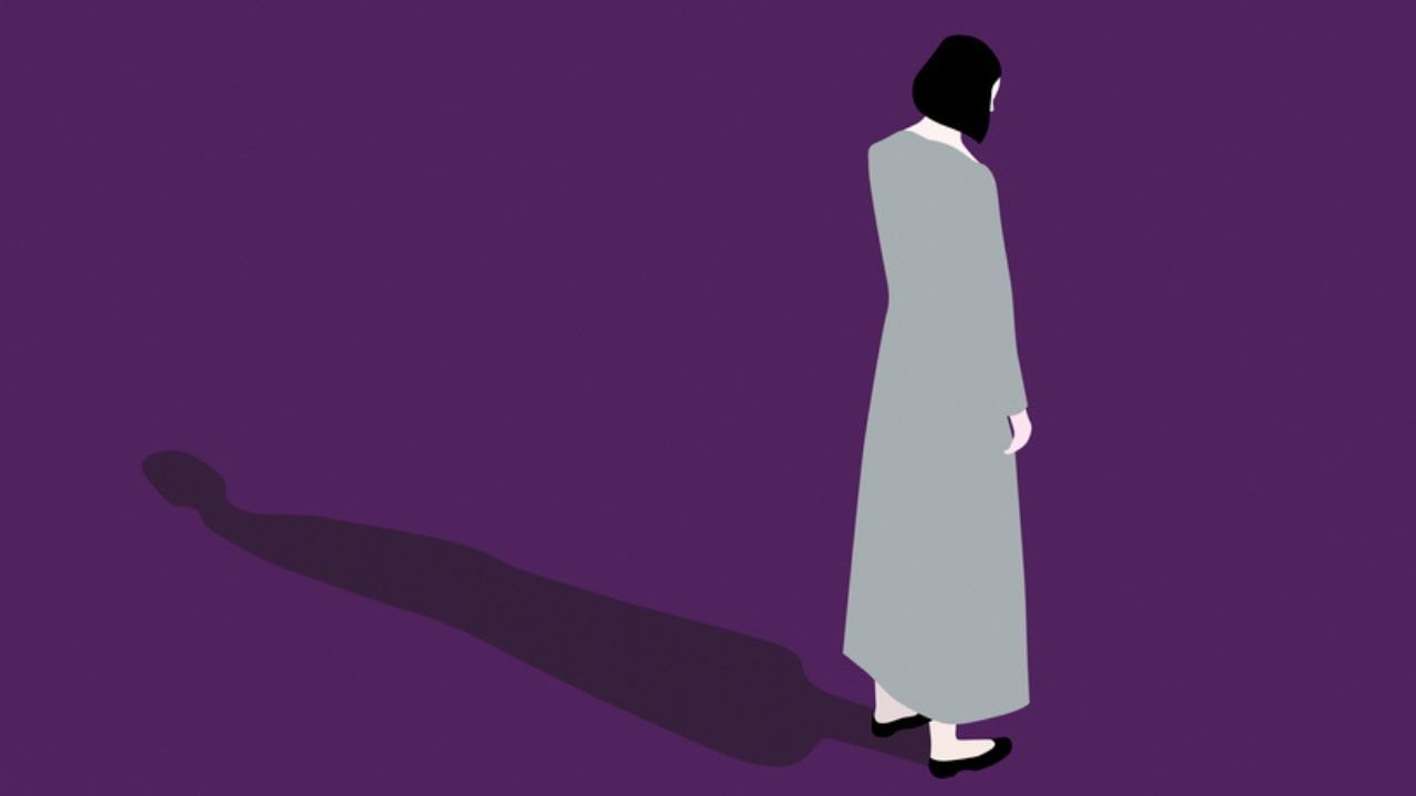 A woman in a long grey dress stands facing away, looking down at the ground, casting a long shadow.