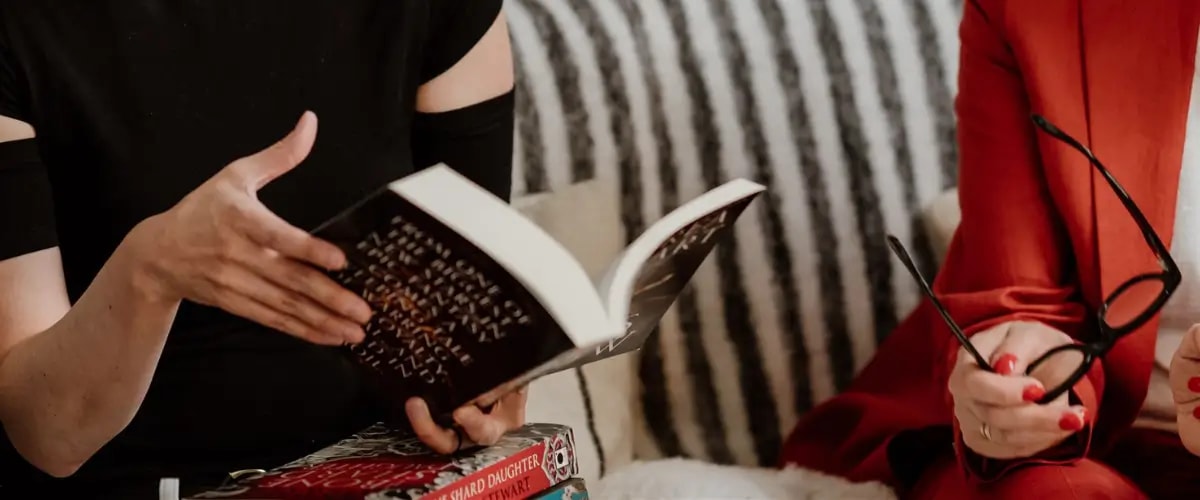 One person flicking through the pages of a book, another beside them holding a pair of glasses.