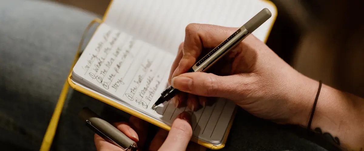 Close-up of a hand writing notes in a yellow notebook with a ballpoint pen.