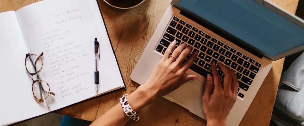 Birds-eye view of a writer typing on their laptop with a notebook, pen, and glasses on the table beside them.