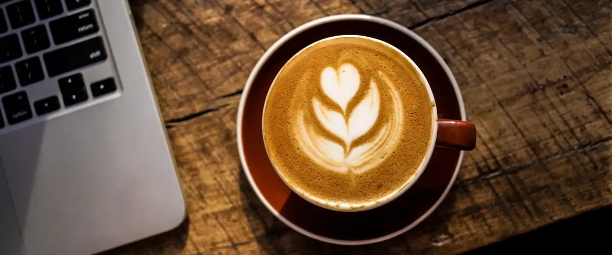 A cup of coffee sits beside a laptop on a wooden table, an intricate heart drawn into the cappuccino foam.