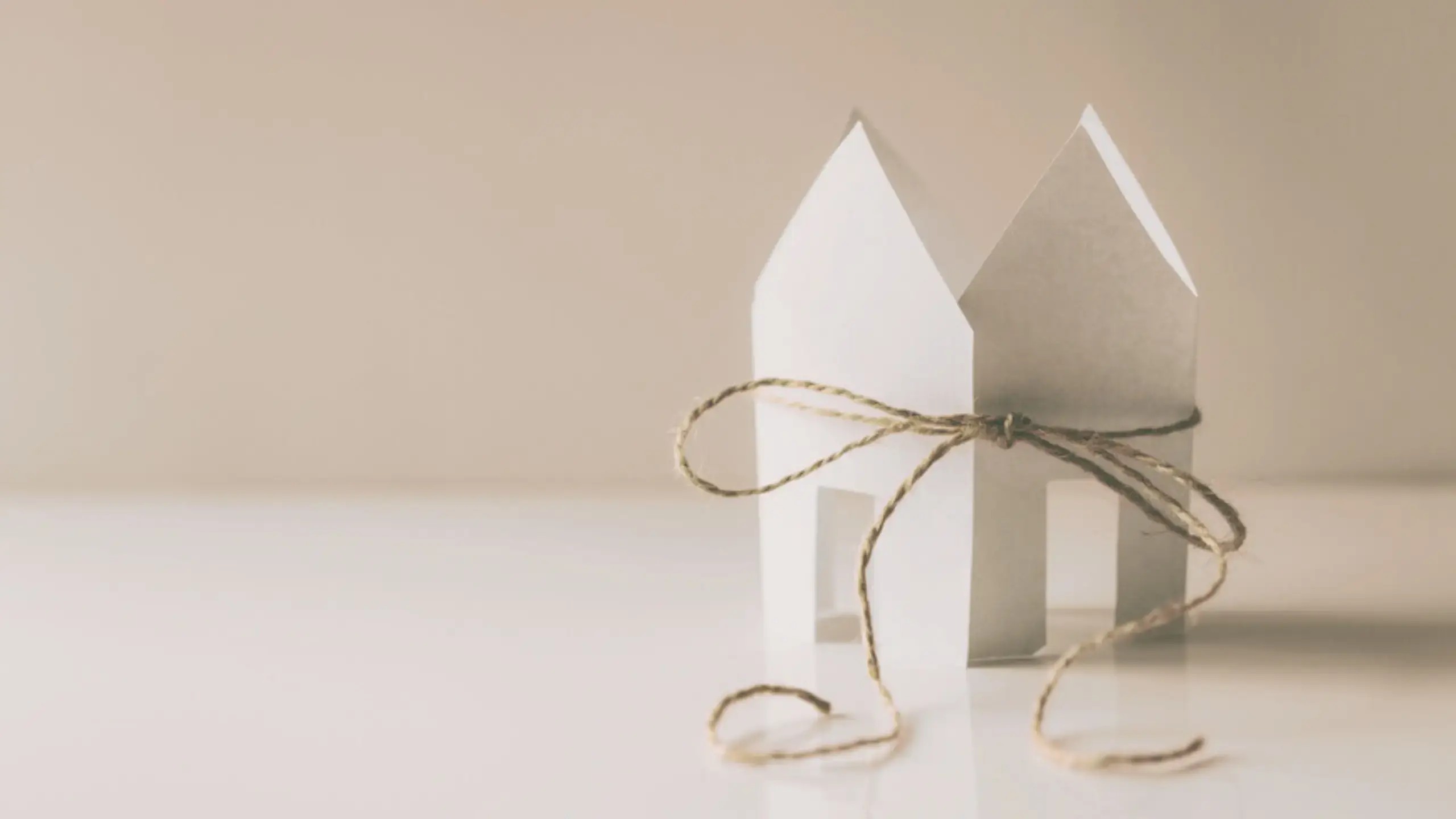 A small house made of folded paper sits on a white surface, held together by a piece of brown string tied in a bow around its middle.