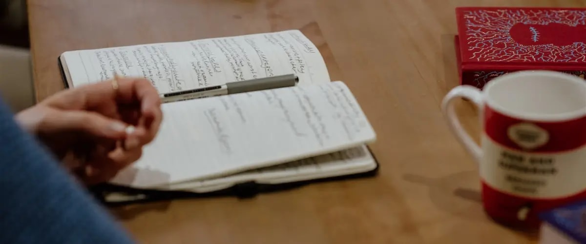 An open notebook filled with handwriting sits on a table, a pen resting on top of it, beside a book, a mug, and a writer's hand.