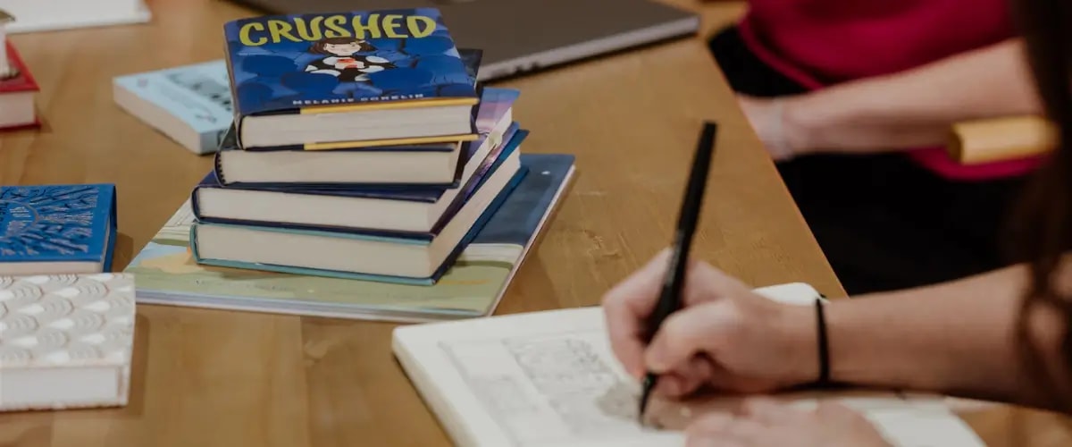 A writer writes by hand in a notebook at a table, with a pile of books stacked next to them, topped by the middle-grade novel Crushed by Melanie Conklin.