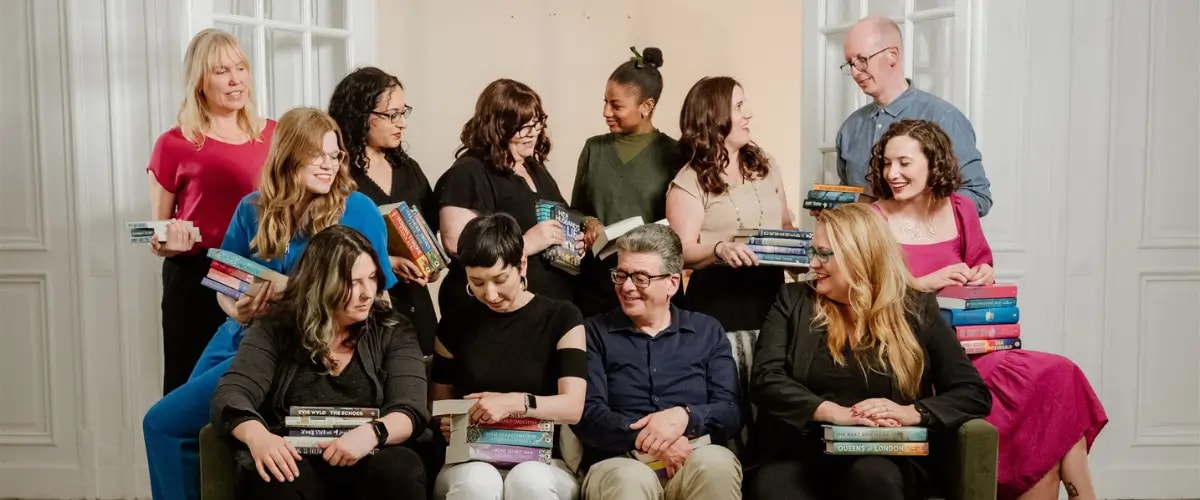 The writing coaches of The Novelry cluster around a sofa with their books, chatting animatedly.
