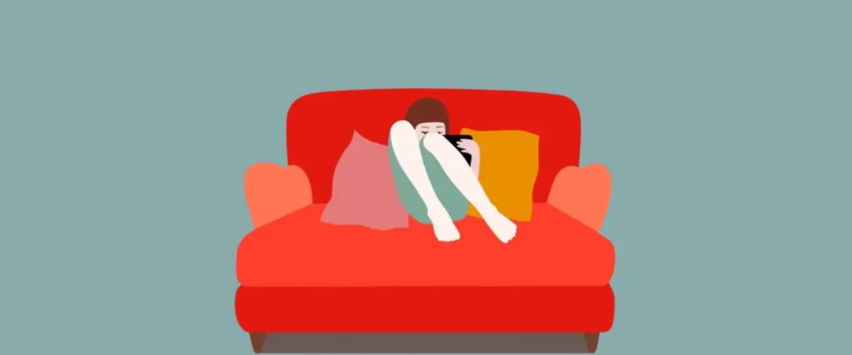 A reader sits in a large red armchair, knees pulled up, fully engrossed in reading a novel.