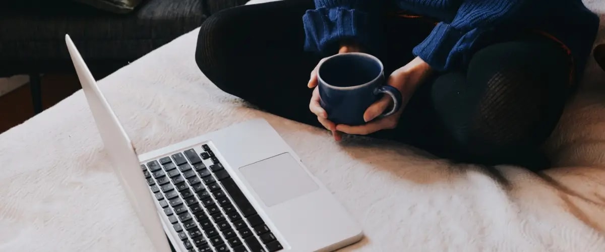 A laptop open on a bed, its owner sitting in front of it with crossed legs and a coffee cup in their hands.