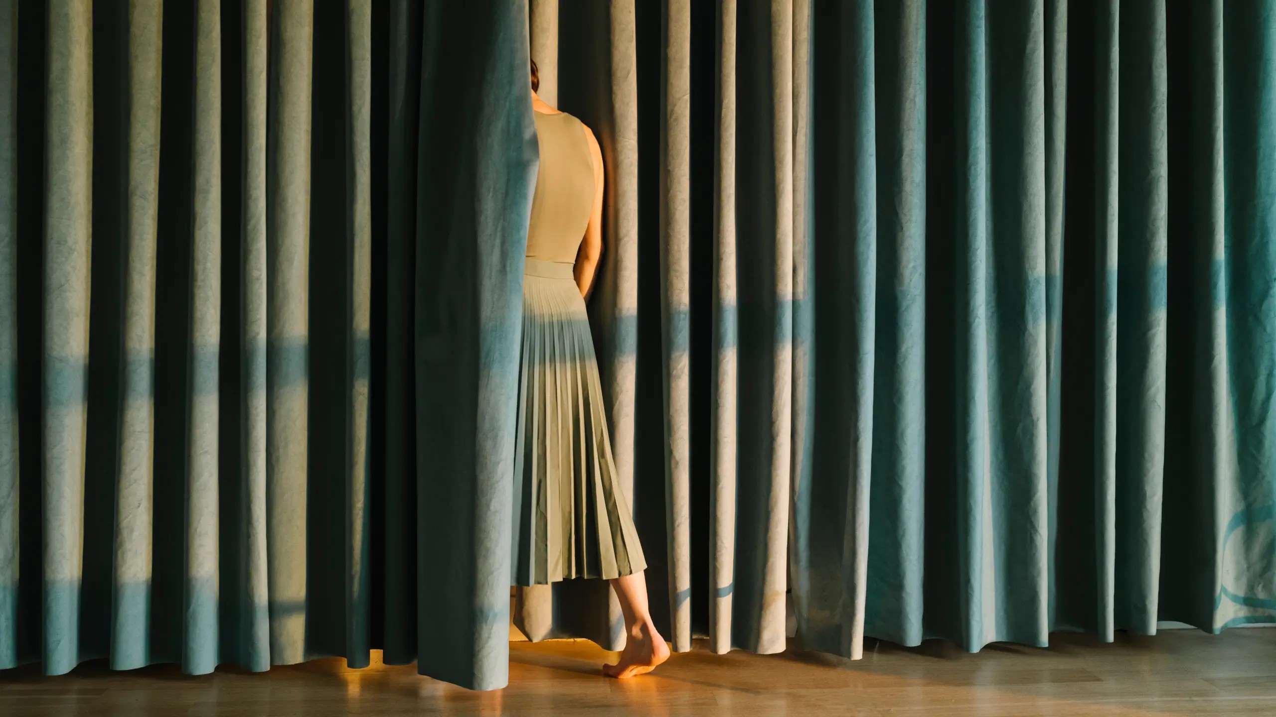 A figure wearing a pleated skirt disappears behind a large velvet curtain.